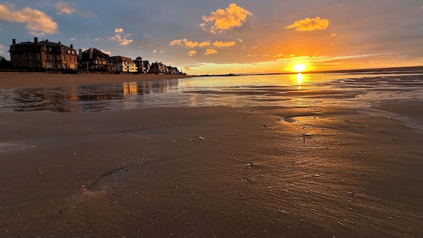 Een strand met een zonsondergang, gebouwen op de achtergrond en een rustige zee.