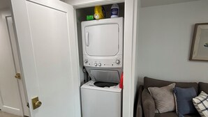 Een kleine wasruimte met een wasmachine en droger, een klein plankje met schoonmaakspullen en een grijze bank met kussens.