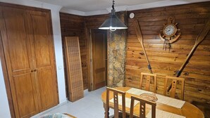Une pièce aux murs en bois, avec un pilier en pierre, une table à manger en bois entourée de chaises, une horloge en bois et une porte en bois.