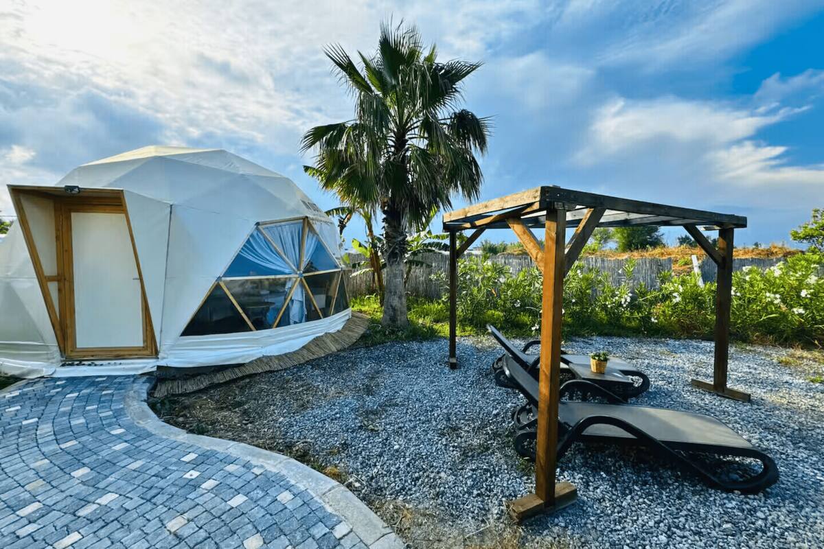 A geodesic dome structure with a wooden door and windows, a wooden pergola, and a gravel path.