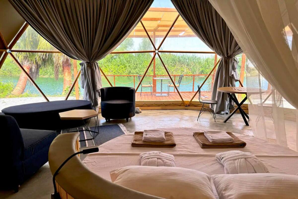 A room with a bed, a chair, a table, and a view of a body of water through large windows.