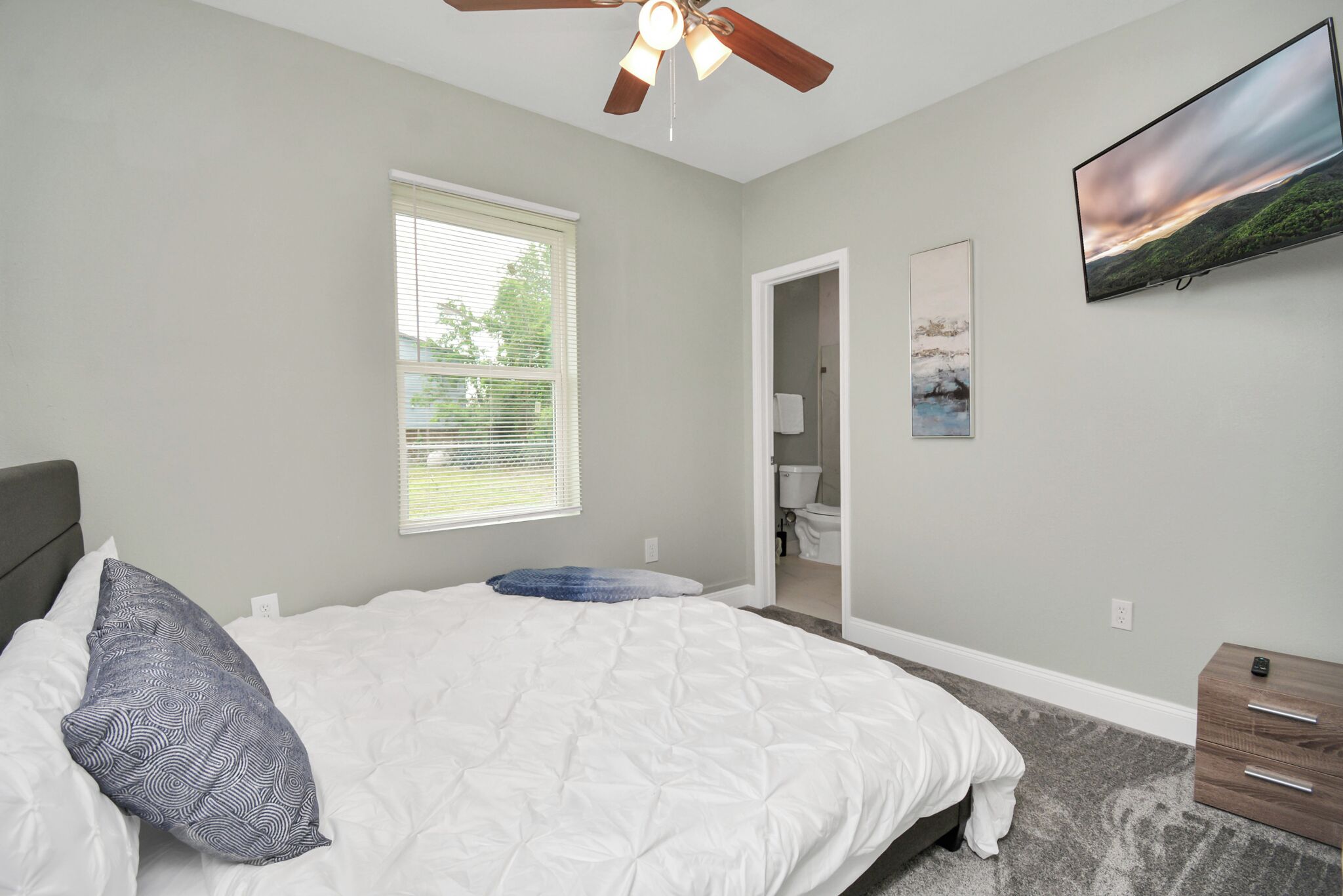 A bedroom with a bed, a ceiling fan, a window, a television, and a door leading to a bathroom.