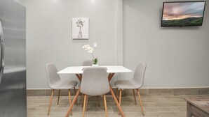 Un espace repas moderne avec une table blanche, quatre chaises grises, un vase avec des fleurs, un tableau encadré accroché au mur et un téléviseur à écran plat fixé au-dessus de la table.