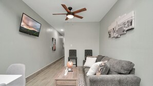 Un salon moderne avec un canapé gris, une table basse, un téléviseur fixé au mur et un ventilateur de plafond.