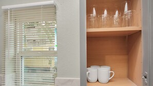 Une salle de bain avec une fenêtre munie de stores, une étagère en bois sur laquelle sont posés des verres et des tasses blanches, et un lavabo blanc.