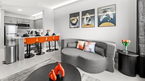 A modern kitchen with grey cabinets, a white countertop, and a stainless steel refrigerator. There is a dining area with orange chairs and a black table. A grey sofa and a black side table with a vase of flowers are in the living area. - Near Disney, Universal, Outlets & OCCC (Orlando)