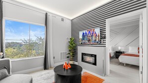 A modern living room with a fireplace, a large window with a view of greenery, and a flat-screen TV mounted on the wall. - Near Disney, Universal, Outlets & OCCC (Orlando)