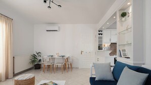 A modern living room with a blue sofa, a dining area with a white table and chairs, and a kitchen area with white cabinets and a glass shelf.