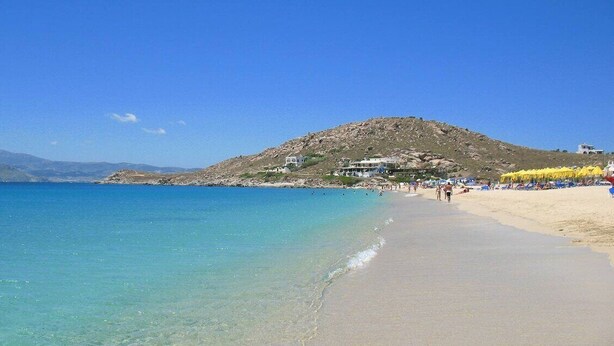 A beach with clear turquoise water, a sandy shore, and a hill with buildings in the background.