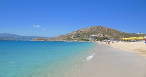 A beach with clear turquoise water, a sandy shore, and a hill with buildings in the background.
