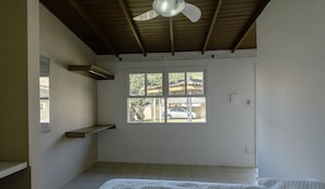 A room with a ceiling fan, a window with a view of a parked car and greenery outside, and a white bed with a white bedspread.