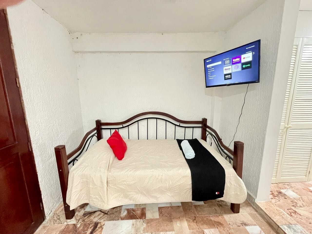 A small, neatly made bed with a red pillow and a black blanket, a wall-mounted TV displaying streaming services, and a wooden door to the left.