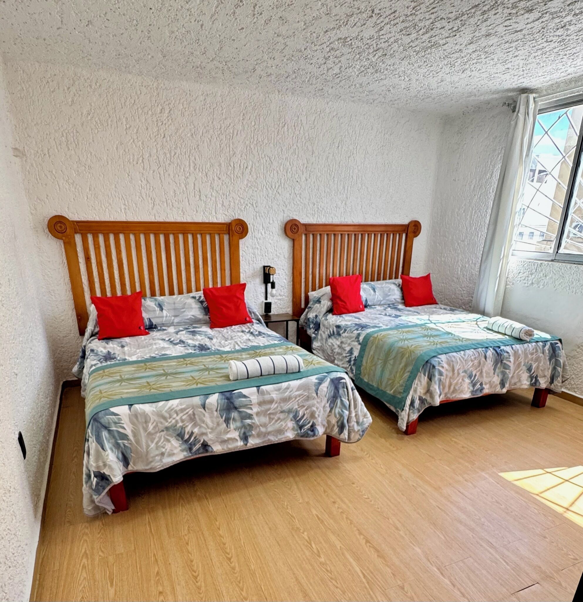 Una habitación con dos camas de madera, cada una con tablero de cabeza con listones, cubiertas con colchas de estampado floral y almohadas rojas. Las paredes son blancas y con textura, y el suelo es de madera.