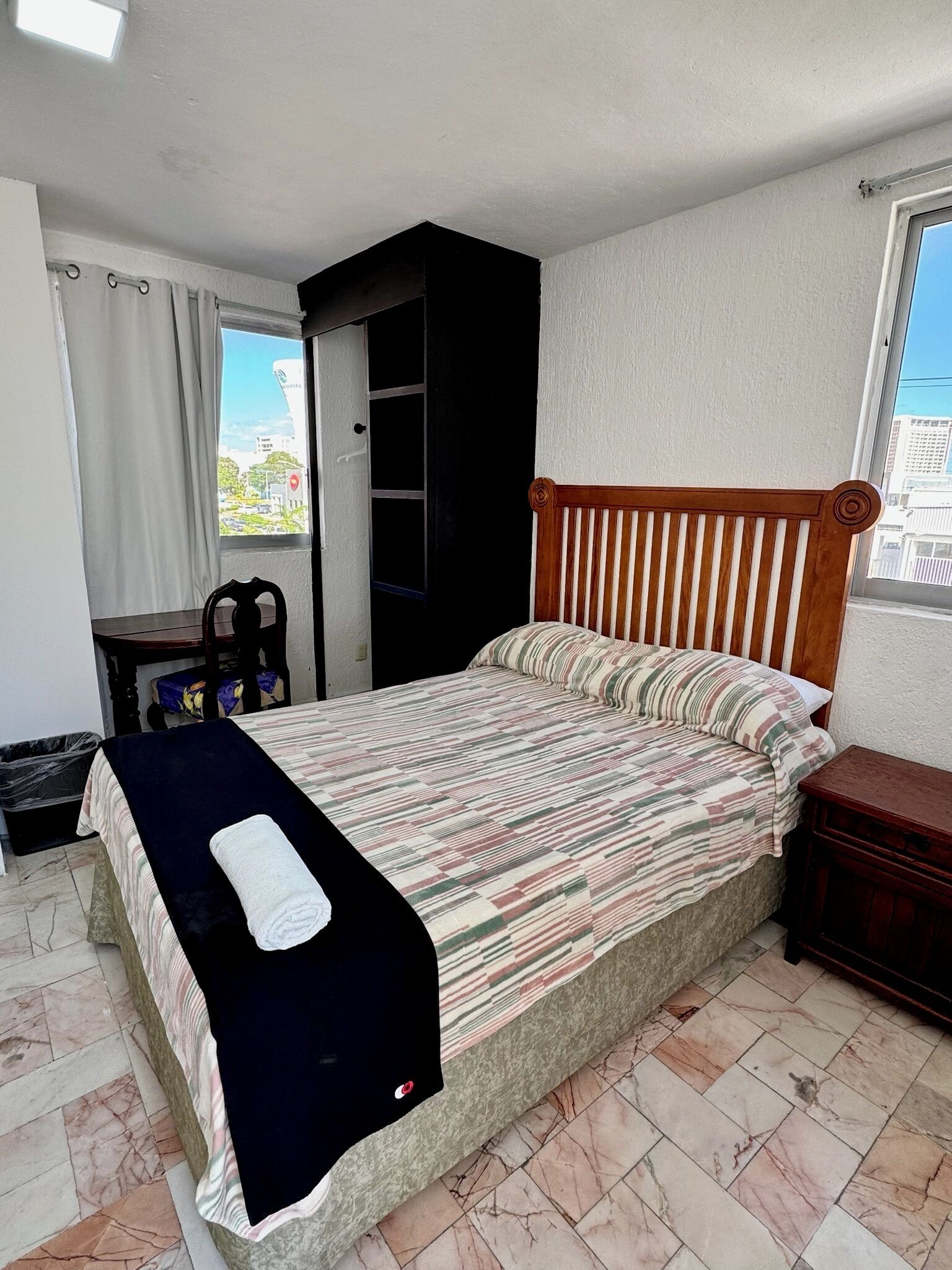 A bedroom with a bed, a wooden headboard, a window with a view, a small table, and a trash bin.