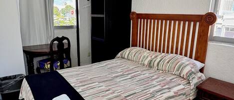 A bedroom with a bed, a wooden headboard, a window with a view, a small table, and a trash bin.