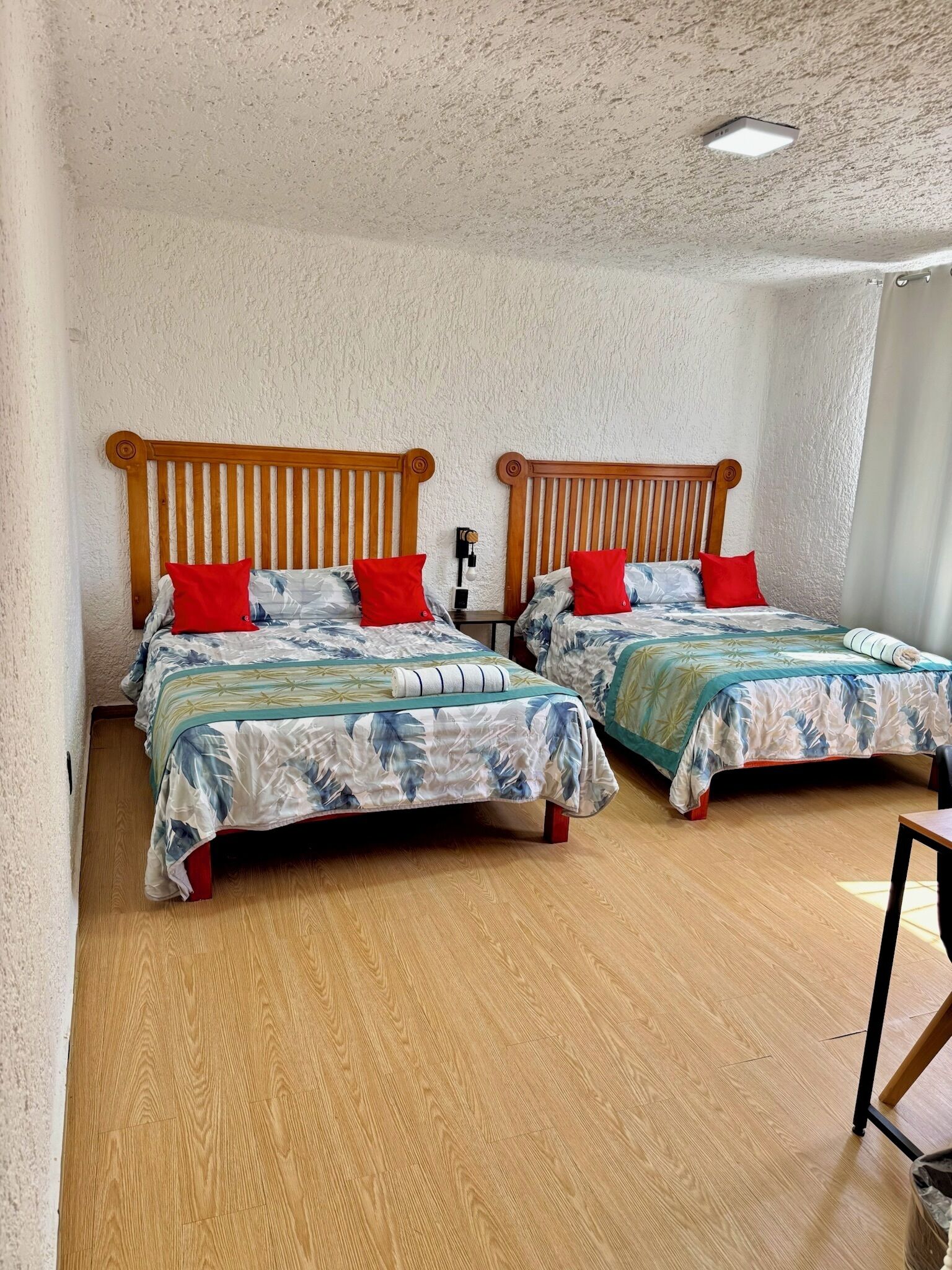 A room with two wooden beds with slatted headboards, each with a red pillow and a floral bedspread. The room has a textured white wall and wooden flooring.