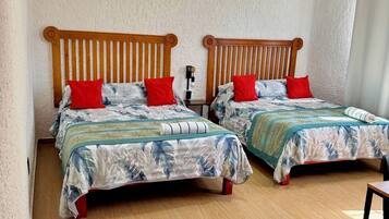 A room with two wooden beds with slatted headboards, each with a red pillow and a floral bedspread. The room has a textured white wall and wooden flooring.