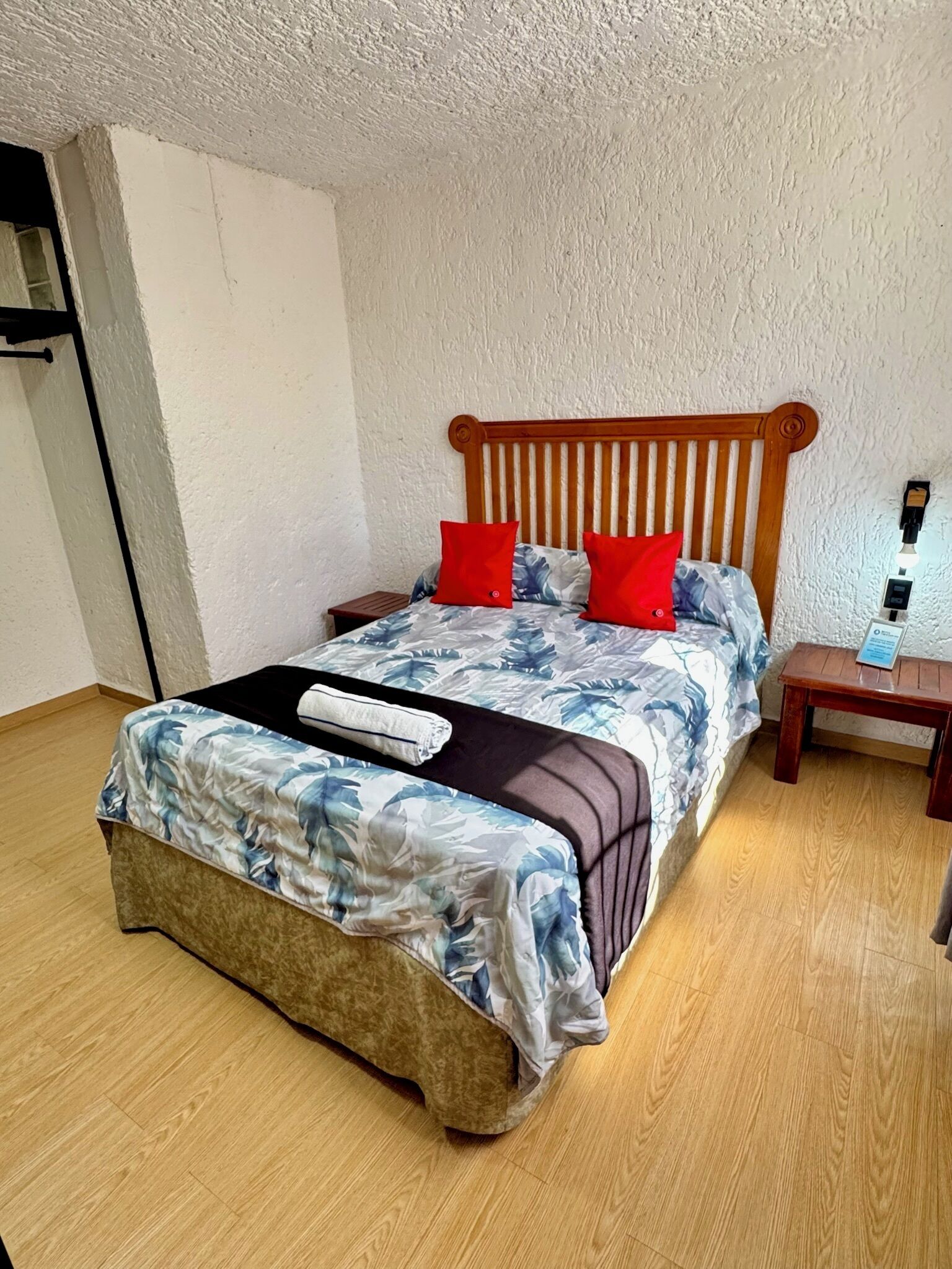A single bed with a wooden headboard, two red pillows, and a floral bedspread. There is a small wooden nightstand beside the bed with a lamp on it. The room has wooden flooring and white walls.