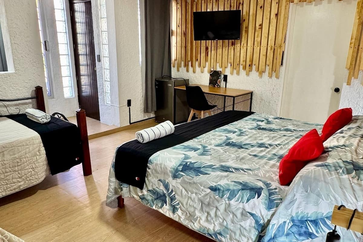 A bedroom with a bed, a desk, a chair, a TV mounted on a wooden wall, and a door leading to a balcony.