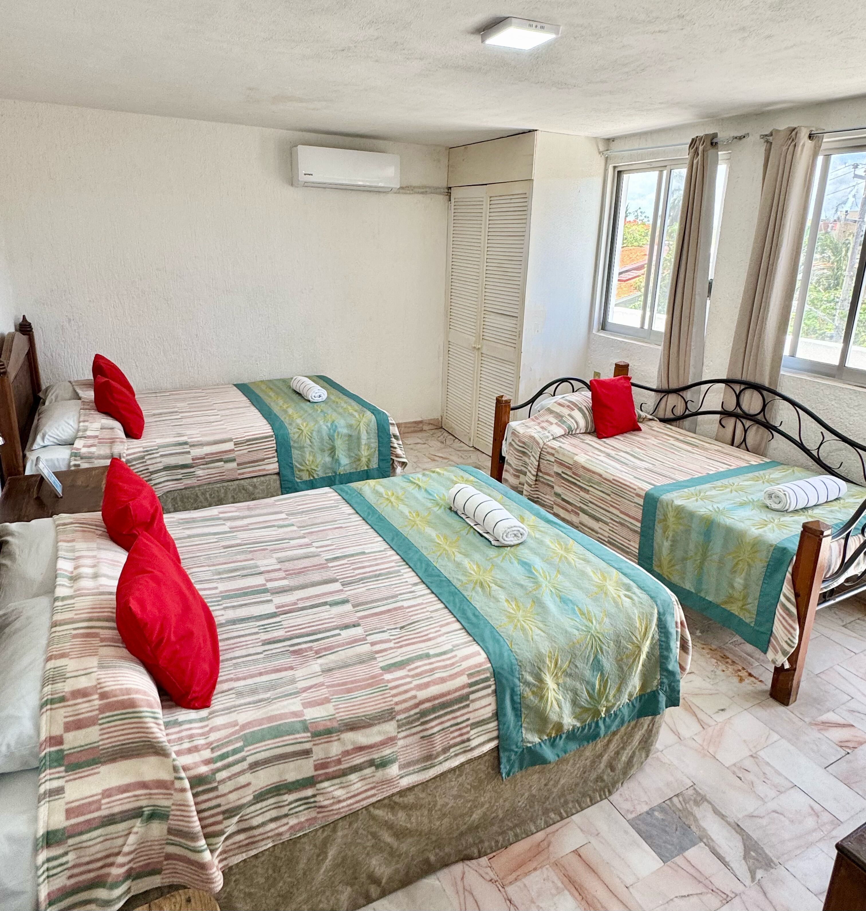 A hotel room with three beds, each with a striped bedspread and red pillows. There is an air conditioning unit on the wall, a window with curtains, and a view of the outside.