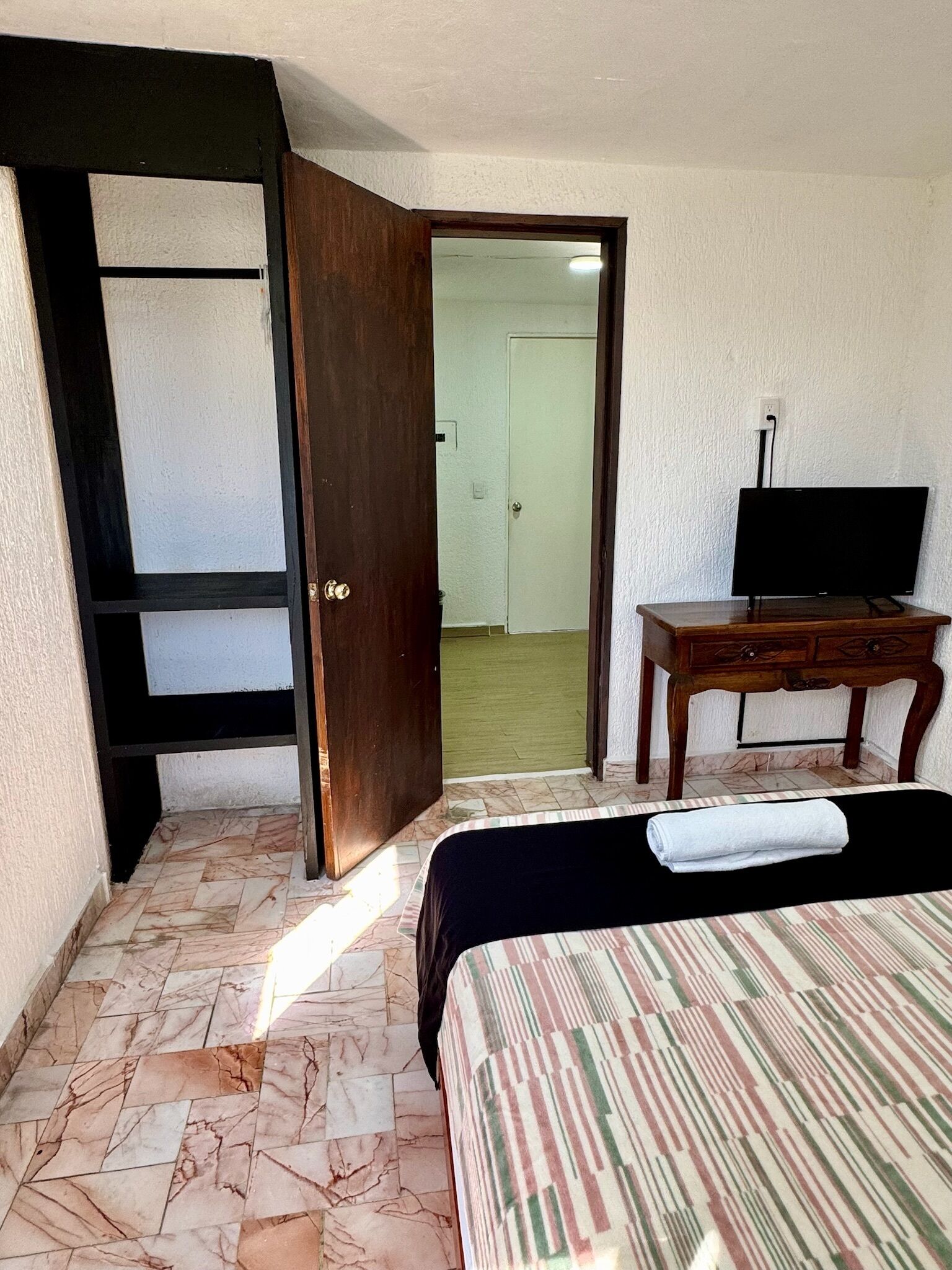 A hotel room with a bed, a desk with a TV, a wardrobe, and a bathroom visible through an open door.