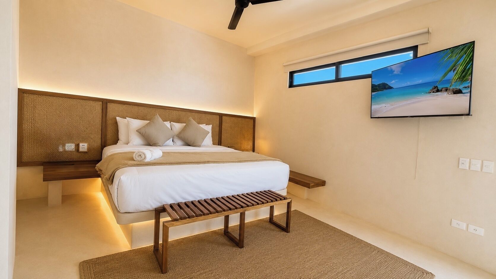 A modern bedroom with a large bed, a wooden bench, a wall-mounted TV, and a window with a beach view.