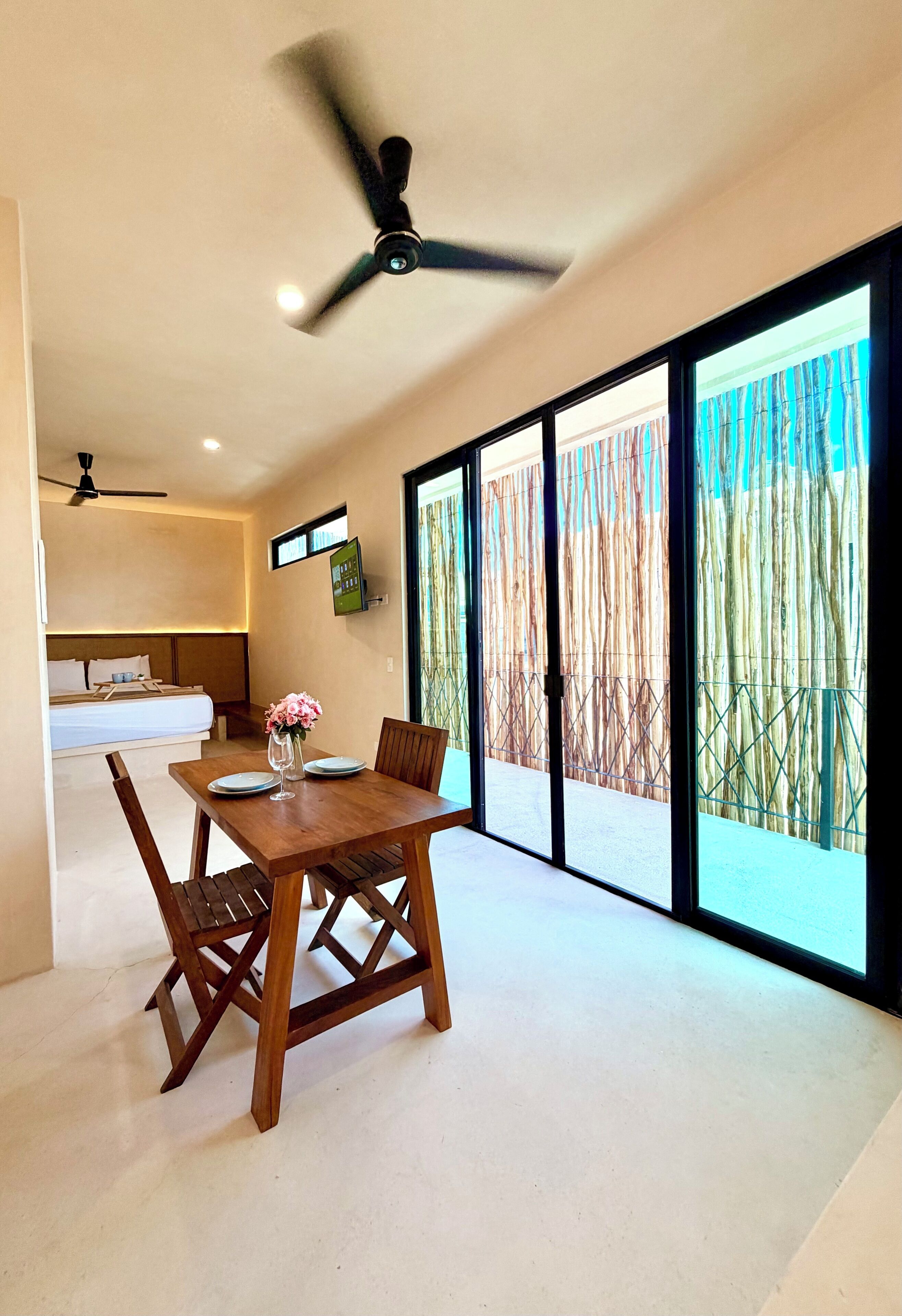 Une chambre d’hôtel moderne dotée d’un coin repas, d’un lit avec une tête de lit, d’un ventilateur de plafond et de grandes portes coulissantes en verre donnant sur une clôture en bambou.