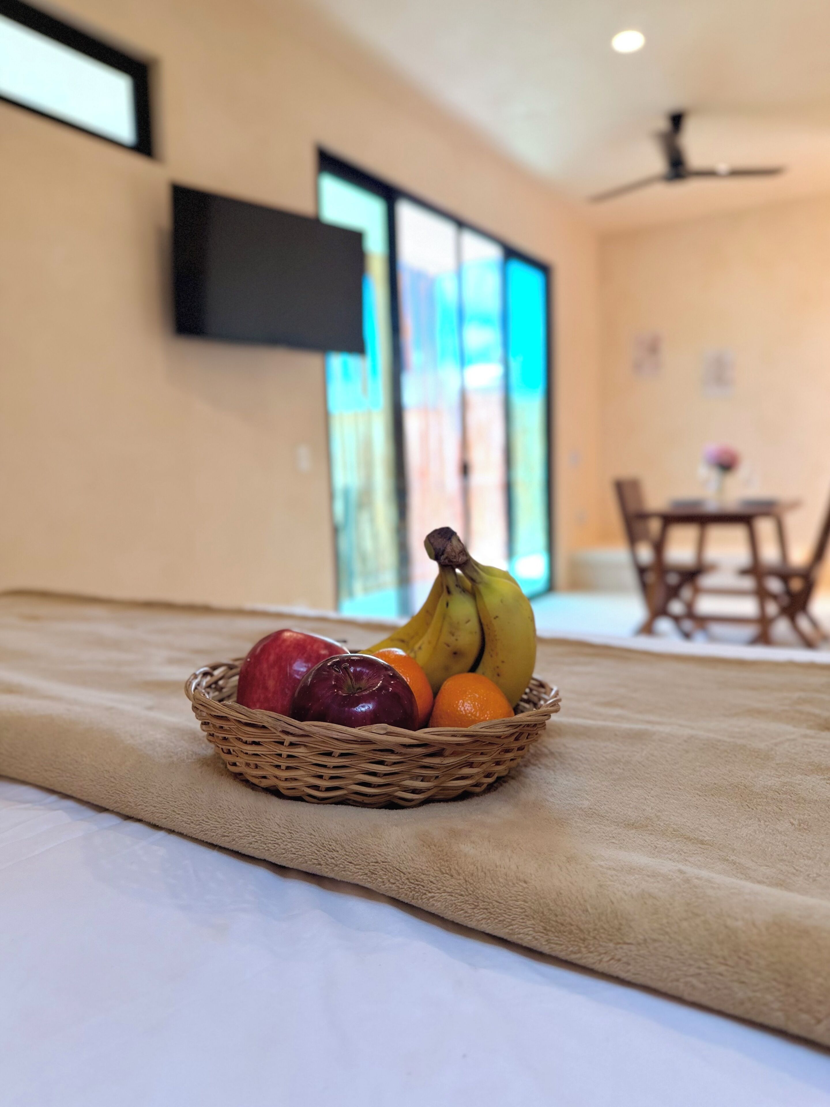 A wicker fruit basket on a bed with a beige blanket, a television on the wall, and a dining area in the background.