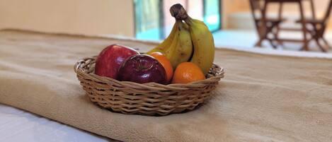 A wicker fruit basket on a bed with a beige blanket, a television on the wall, and a dining area in the background.