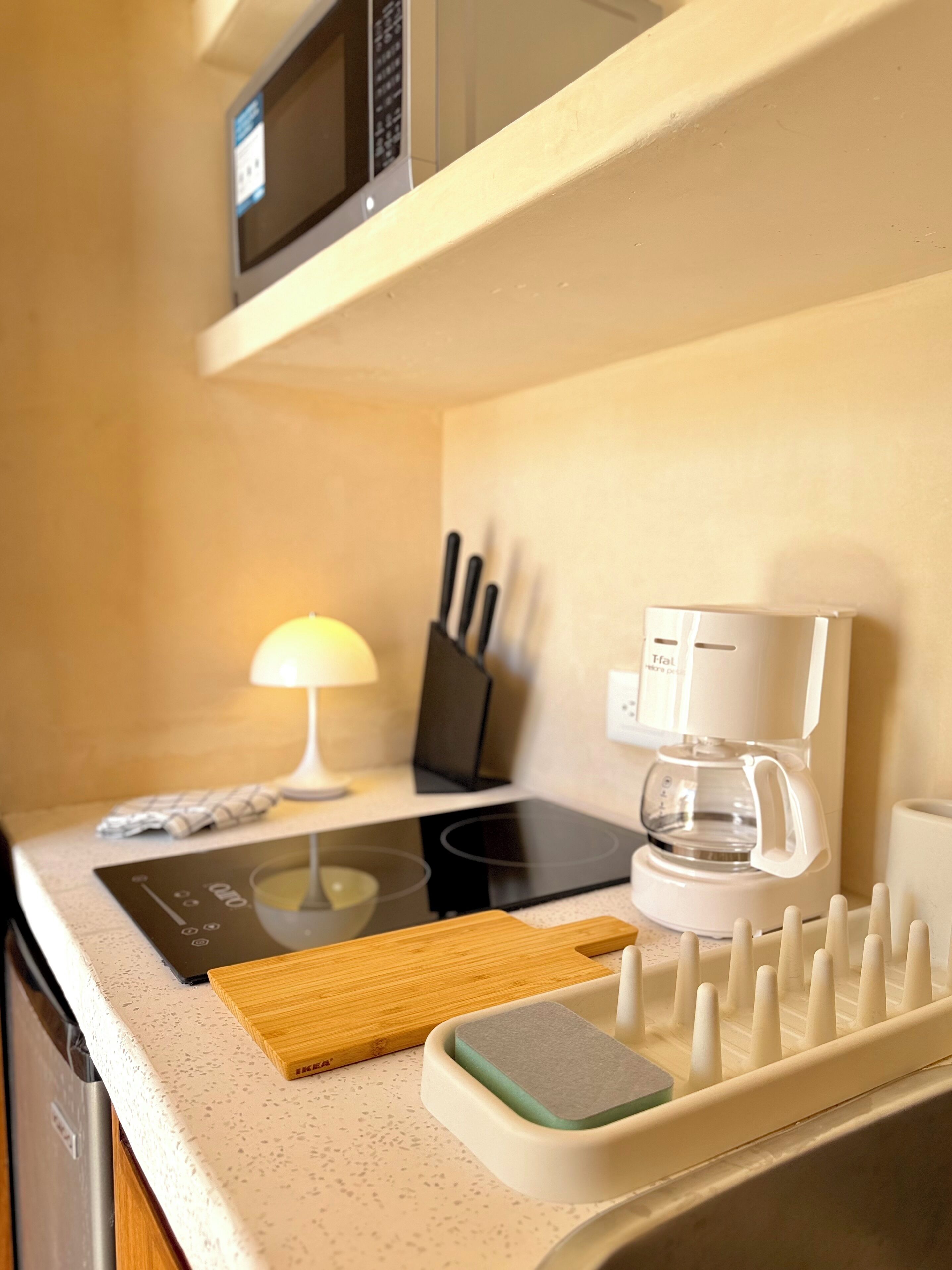 Une cuisine équipée d’un four à micro-ondes, d’une machine à café, d’une plaque de cuisson à induction et d’une planche à découper.