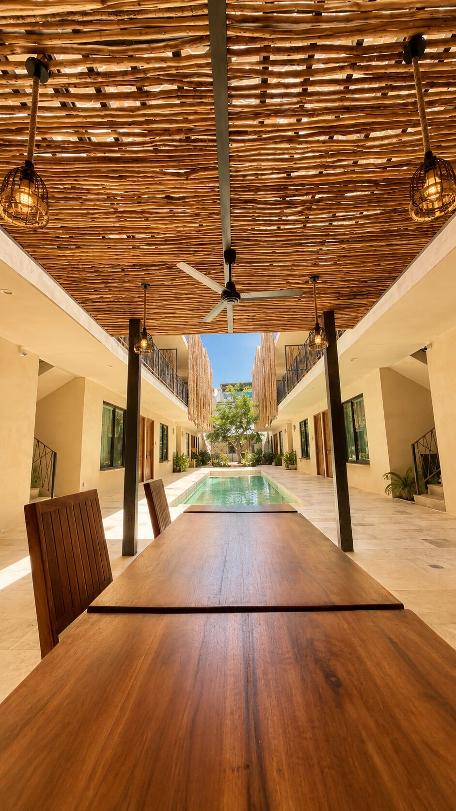 A spacious indoor area with a long wooden table, a ceiling fan, and a view of a pool and greenery outside.