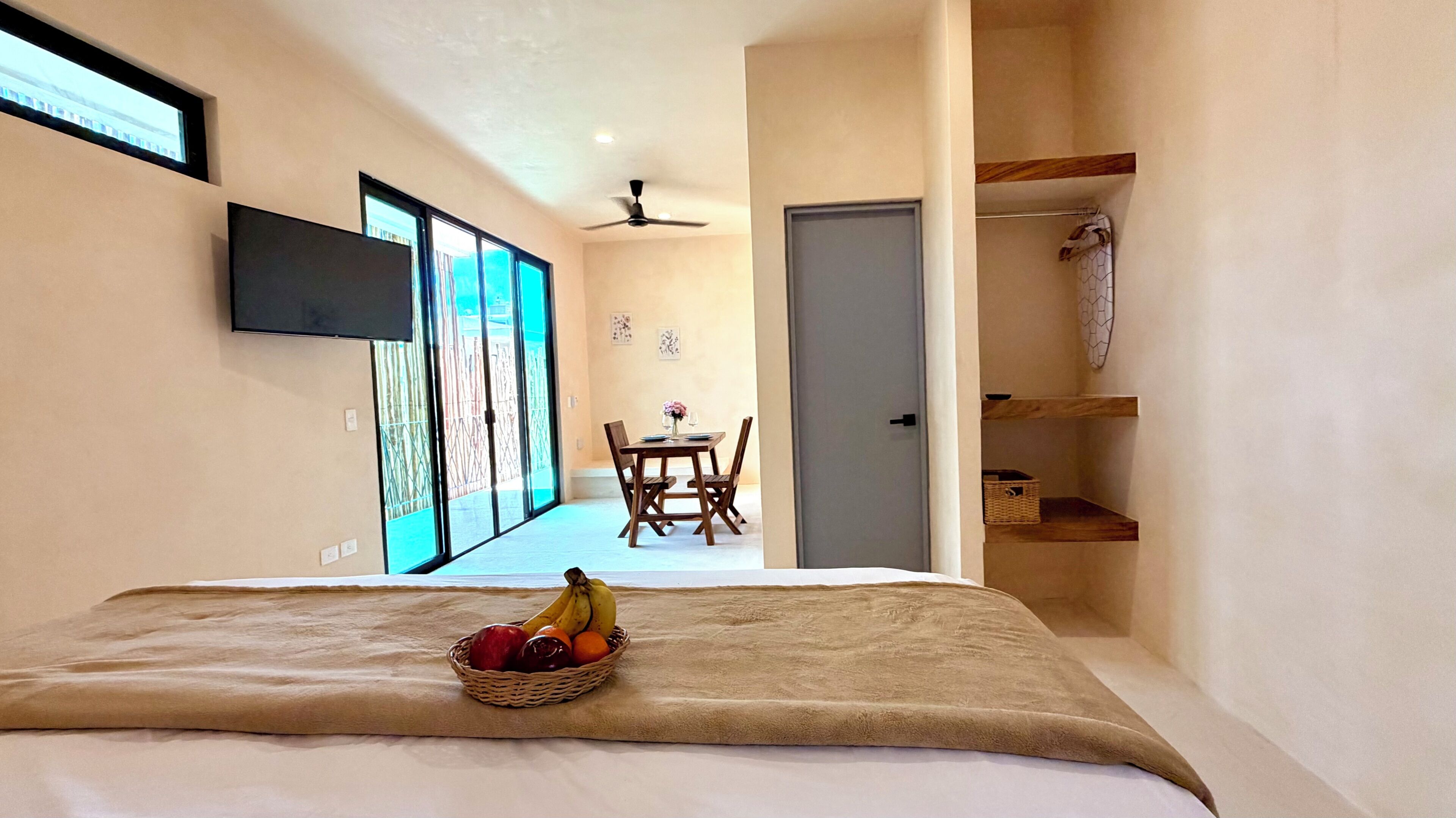 Quarto com cama, cesta de frutas, área de refeições com mesa e cadeiras, e televisão na parede.