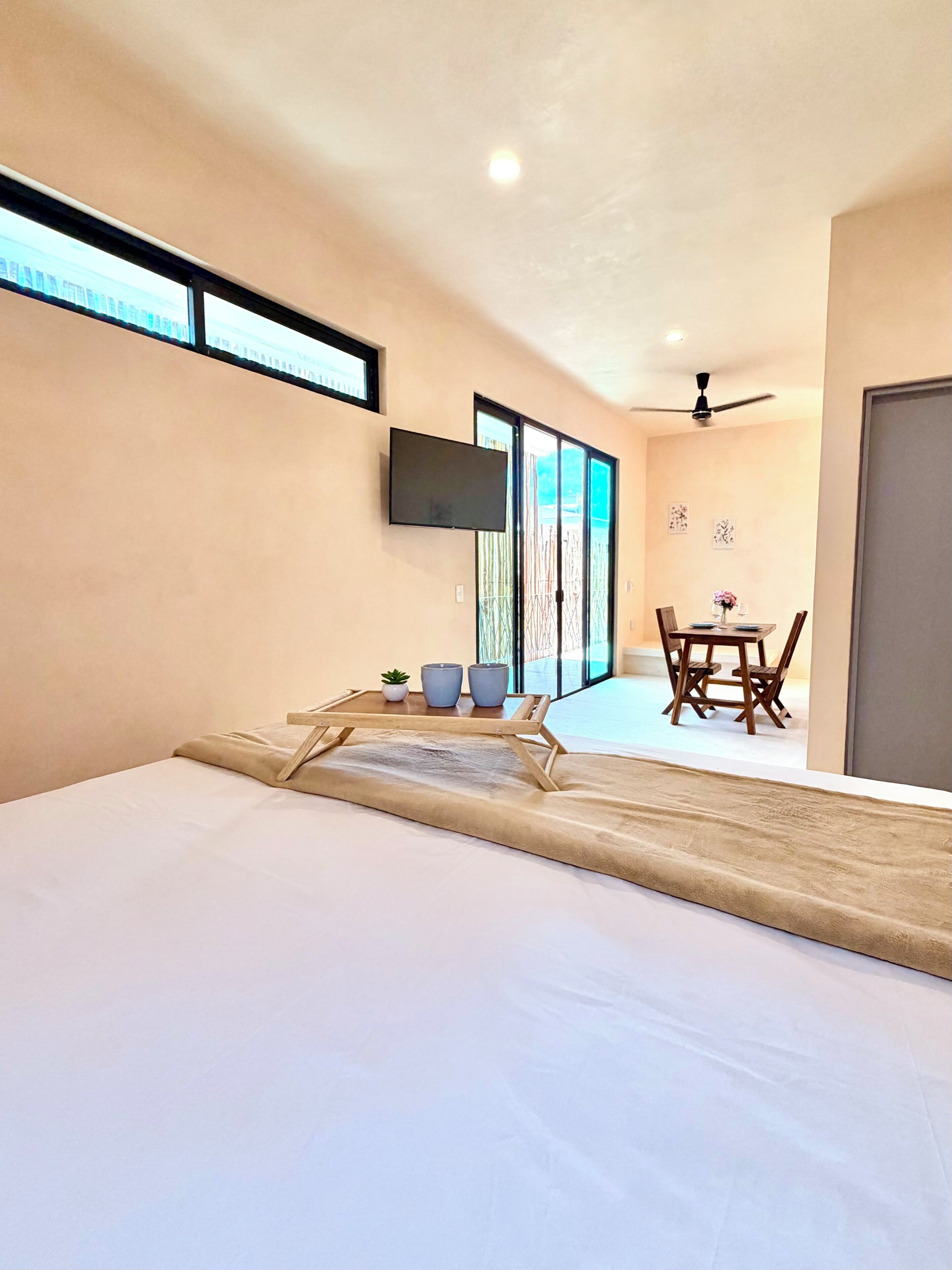 A modern hotel room with a bed, a TV, a ceiling fan, and a dining area with a table and chairs.