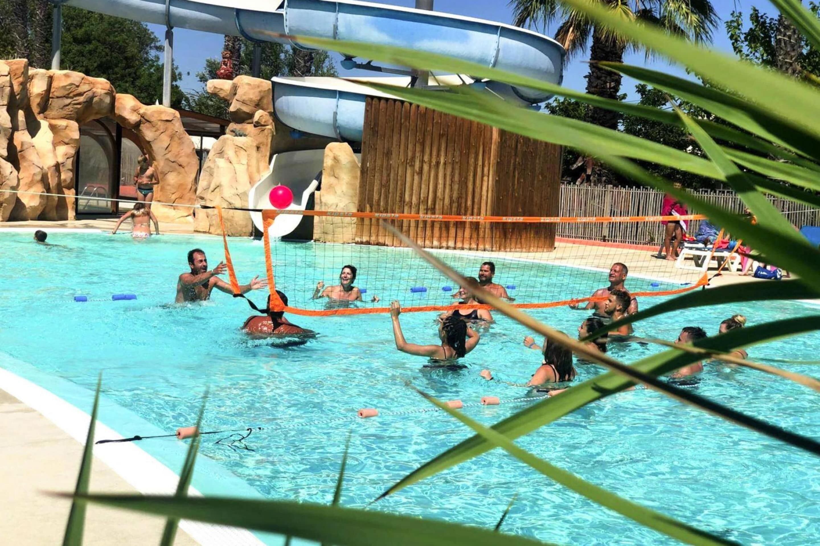 A swimming pool with a volleyball net in the center, surrounded by palm trees and a water slide in the background.