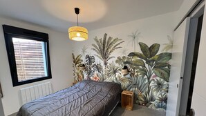 Une chambre avec un lit, une fenêtre avec des stores, une fresque murale représentant des plantes tropicales, un luminaire suspendu et un radiateur.