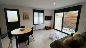 Un salon moderne comprenant un canapé, une table à manger avec des chaises, un téléviseur fixé au mur et une porte-fenêtre coulissante donnant sur un espace extérieur avec une table et des chaises.