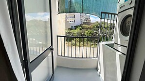 Un balcón con lavadora, un ventanal pequeño y vistas a edificios y vegetación.