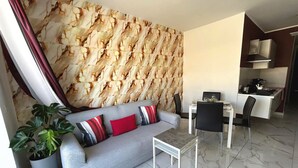 Una sala de estar moderna con una pared con diseño de mármol, un sofá gris con cojines rojo y negro, una mesita blanca pequeña y una planta en maceta.