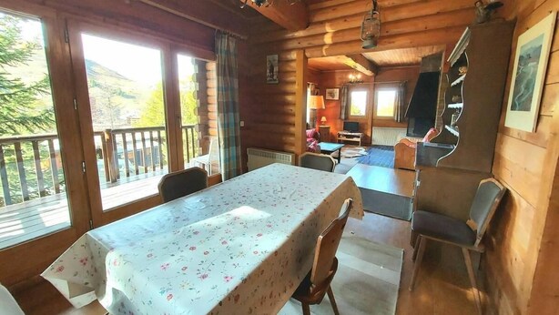 Interno accogliente di una baita con tavolo da pranzo, sedie, angolo cottura e un balcone con vista sul paesaggio.