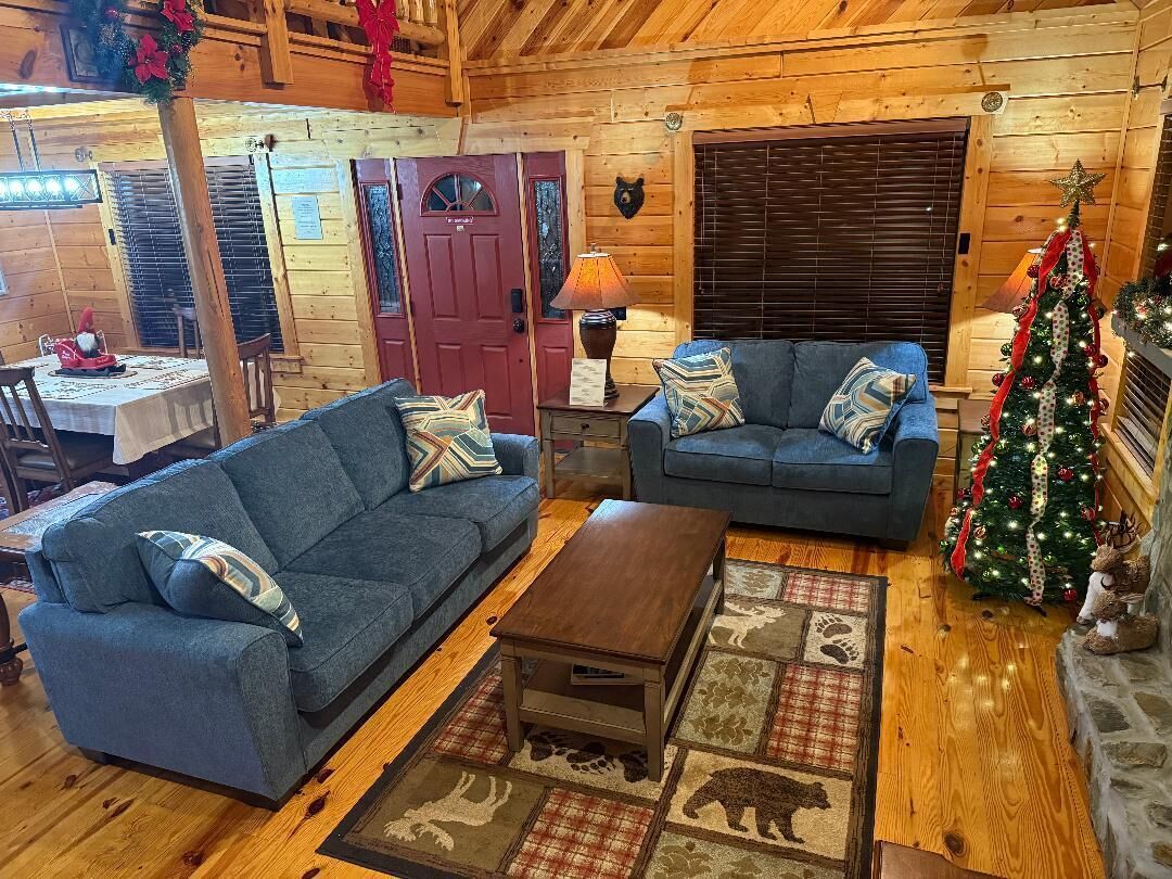 A cozy log cabin interior with two sofas, a wooden coffee table, a Christmas tree, and a fireplace.