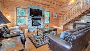 A cozy log cabin interior with a stone fireplace, leather furniture, and a wooden staircase.
