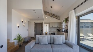 A modern living room with a grey sofa, a staircase, a kitchen, and a balcony with lounge chairs.