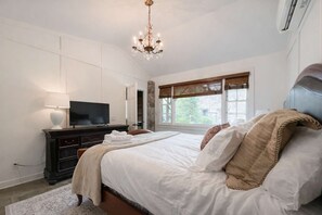 Een slaapkamer met een bed, een televisie op een dressoir en een raam met jaloezieën.