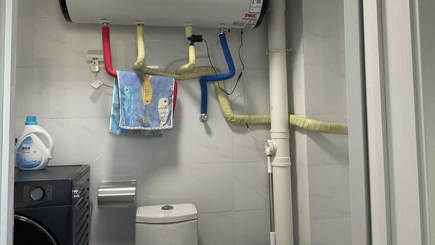 Une salle de bain avec des toilettes blanches, un lave-linge et un chauffe-eau doté de plusieurs tuyaux.