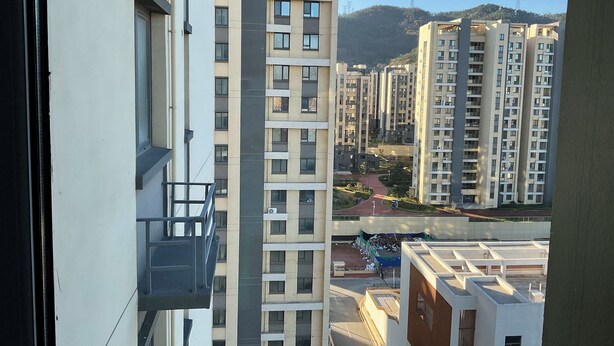 A high-rise residential building with multiple floors, balconies, and a view of a hilly landscape in the background.