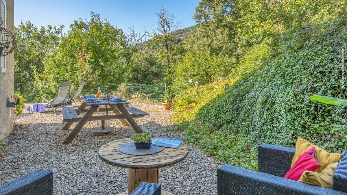 An outdoor seating area with a wooden table and chairs, surrounded by greenery and a fence.