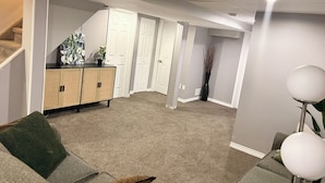 Una sala de estar con un sofá gris, una mesita y una lámpara. Hay dos puertas y una escalera que sube a un nivel superior.