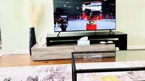 Un salón con televisor de pantalla plana sobre un soporte de madera, un reloj en la pared y un florero con una pluma.