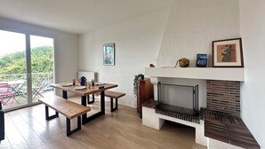 A modern living room with a fireplace, a dining area with a wooden table and benches, and a balcony with a view of greenery.