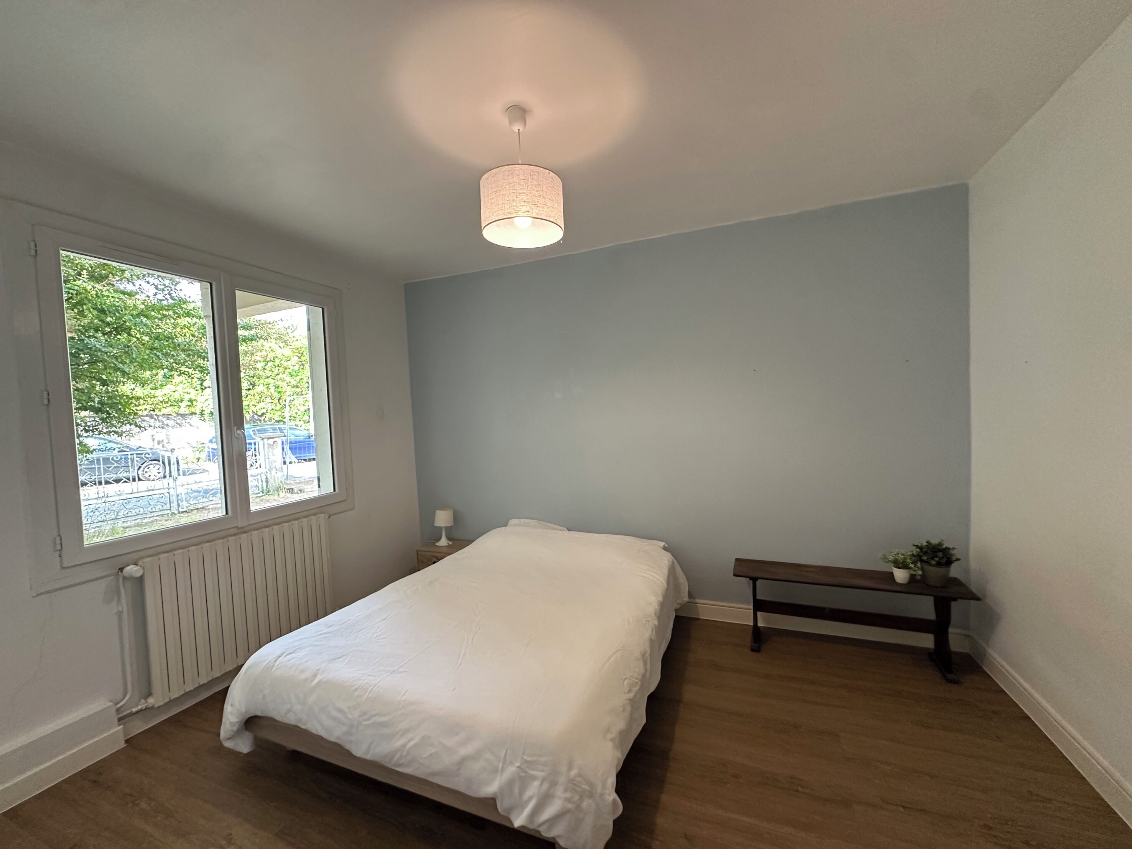 A bedroom with a bed, a window, a radiator, a bench, and a hanging light fixture.